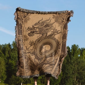 May include: A brown woven throw blanket featuring a detailed illustration of a dragon. The dragon is surrounded by trees and foliage. The blanket has a fringed edge and is held up against a blue sky background.