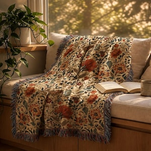May include: A woven throw blanket featuring a detailed floral pattern with orange, red, and green botanical motifs on a cream background. The rectangular blanket has fringed edges along the sides and is draped over a cushioned seat with an open book.