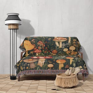 May include: A woven throw blanket with a mushroom and floral pattern in shades of red, yellow, brown, and green. The blanket is draped over a couch with a woven basket and a floor lamp in the background.