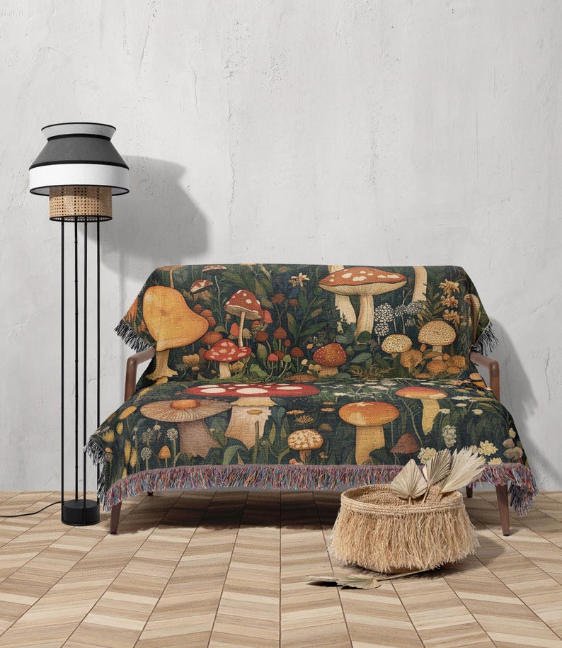 May include: A woven throw blanket with a mushroom and floral pattern in shades of red, yellow, brown, and green. The blanket is draped over a couch with a woven basket and a floor lamp in the background.