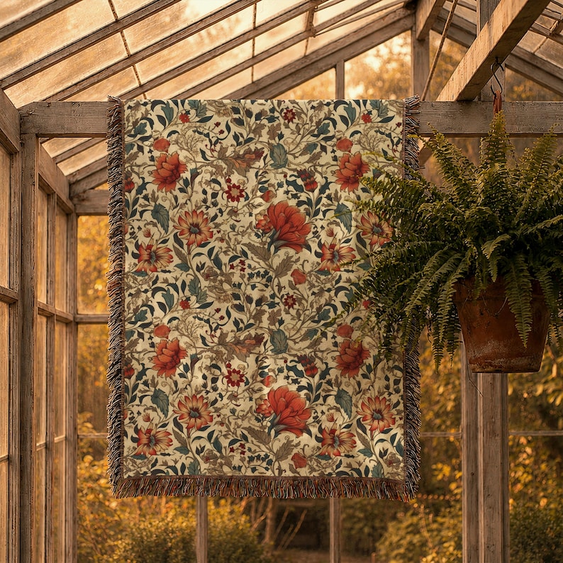 May include: A woven throw blanket featuring a repeating botanical pattern of large red flowers, smaller dark red blossoms, and intricate green foliage on a cream-colored background, finished with a fringed edge along the top and bottom sides.