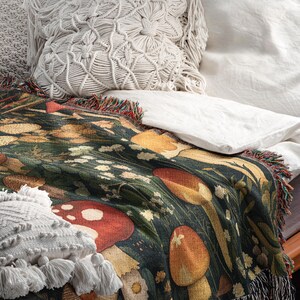 May include: A woven tapestry throw blanket with a dark green background and a colorful mushroom design. The blanket has fringe on all sides and is draped over a couch with white pillows.