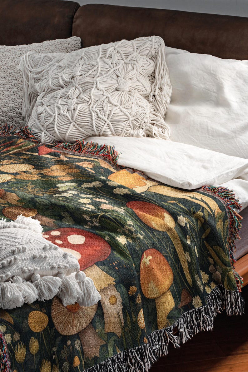 May include: A woven tapestry throw blanket with a dark green background and a colorful mushroom design. The blanket has fringe on all sides and is draped over a couch with white pillows.
