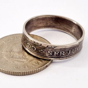 May include: A silver ring made from a coin with the inscription "SFR JUGOSLAVIJA" on the outside of the band. The coin is lying on its side next to the ring.
