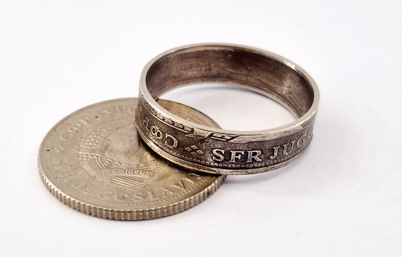 May include: A silver ring made from a coin with the inscription "SFR JUGOSLAVIJA" on the outside of the band. The coin is lying on its side next to the ring.