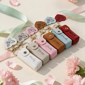 May include: A collection of personalized keychains with heart-shaped pendants featuring butterfly designs. Each keychain has a rectangular case with a name printed on it. The keychains are in various colors, including white, beige, pink, brown, blue, and red.