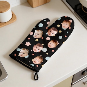 May include: Black oven mitt featuring multiple baby faces, along with pastel-colored polka dots and star shapes. The mitt has a black trim and a loop for hanging. A unique kitchen accessory.