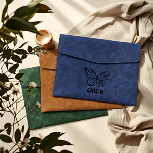 May include: Three leather-like document holders in blue, brown, and green. The blue holder features a butterfly and floral design with the name "Chloe" printed below. The holders have a flap closure and are stacked on top of each other.