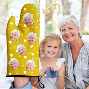 May include: Yellow oven glove with a repeating pattern of smiling faces and white hearts. The glove is being worn by a person. A woman and a young girl are in the background.
