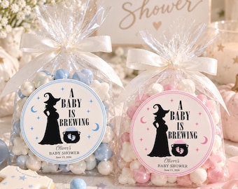 May include: Two clear cellophane bags filled with pastel candies, tied with white satin ribbons. Each bag features a circular label with a black silhouette of a pregnant witch, a cauldron, and the text A BABY IS BREWING, Oliver's BABY SHOWER, June 13, 2026.