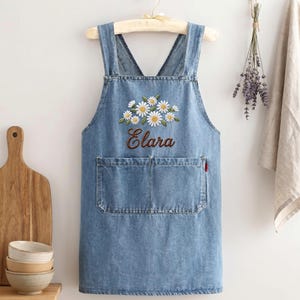 May include: A denim apron with a large front pocket and adjustable straps. The apron features an embroidered design of daisies and the name "Clara" in a cursive font. The apron is hanging on a wooden hanger.