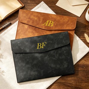 May include: Two rectangular document folders with envelope-style flaps. One folder is tan with the gold embossed letters AB, and the other is black with the gold embossed letters BF. Both folders feature a smooth, matte finish and clean edges.