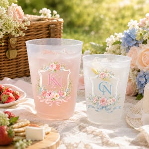 May include: Two clear plastic cups filled with ice and liquid. The larger cup features a pink floral crest with the monogram KN, while the smaller cup displays a blue floral crest with the monogram CN. Both cups have a decorative ribbon design.