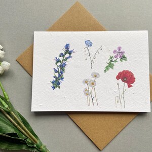 May include: A handmade greeting card with watercolour illustrations of wildflowers, including bluebells, daisies, and a red poppy. The card rests on a brown envelope, with a sprig of lily of the valley in the corner.