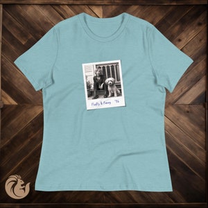 May include: Light blue t-shirt featuring a black and white Polaroid-style image. The image shows a person and a dog sitting on a porch, with the text "Fluffy & Penny '96". The shirt is made of soft material.