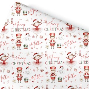 May include: White wrapping paper featuring a festive pattern of Christmas illustrations. The design includes ballerinas, toy soldiers, snow globes, and the text "Merry Christmas" in red. The name "Millie" is also written in red.