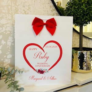 May include: A white gift bag featuring a red satin bow at the top. The centre displays a red heart graphic with the text "HAPPY RUBY WEDDING ANNIVERSARY" and the date "05.06.2026". Below the heart, the names "Margaret & Arthur" are printed in red.
