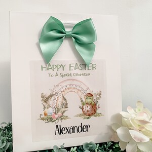 May include: White gift bag with a green bow. The bag has a sticker with a rainbow, a green dinosaur, and the text "Happy Easter To A Special Grandson Alexander".