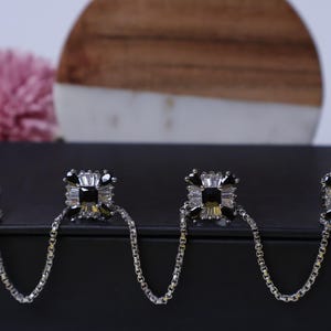 May include: Four silver-toned square earrings with black and clear crystal accents, connected by delicate chains. The earrings are displayed on a black surface, with a blurred pink flower and a wooden and white background.