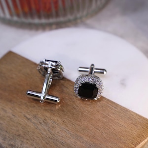 May include: A pair of silver cufflinks with black square stones surrounded by clear rhinestones.