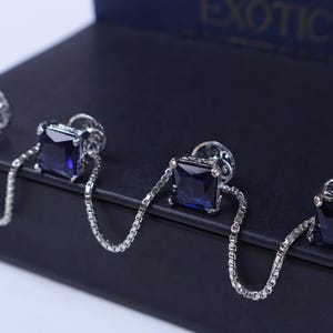 Puede incluir: Juego de cuatro gemelos plateados con piedras cuadradas de color azul oscuro. Cada gemelo está conectado por una delicada cadena. Los gemelos se muestran sobre una superficie oscura y texturizada, con la palabra "EXOTIC" visible.