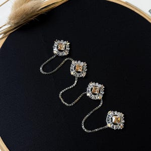 Puede incluir: Cuatro broches de cadena de plata y pedrería con forma cuadrada y una piedra de color claro y ámbar en el centro. Los broches están dispuestos en fila sobre un fondo negro.