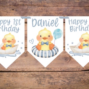 May include: A banner set with a watercolor illustration of a yellow duckling wearing a blue bow tie. The banner includes the text "Happy 1st Birthday" and the name "Daniel". The duckling is in a paper boat and a striped inner tube, holding a blue balloon.