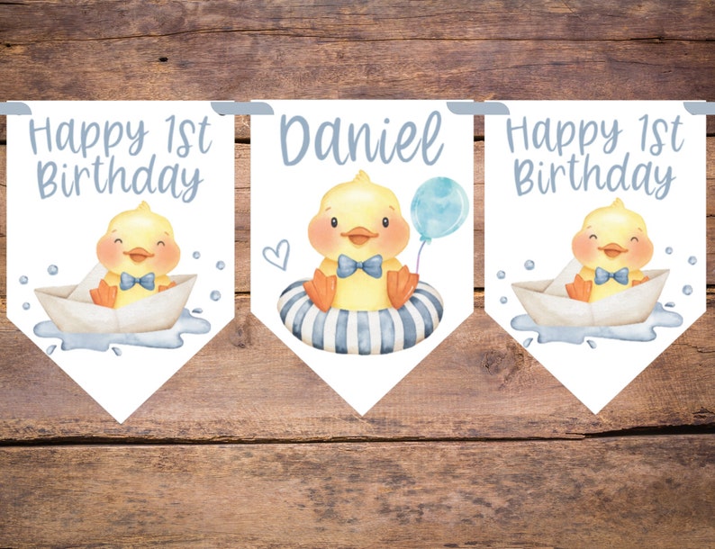 May include: A banner set with a watercolor illustration of a yellow duckling wearing a blue bow tie. The banner includes the text "Happy 1st Birthday" and the name "Daniel". The duckling is in a paper boat and a striped inner tube, holding a blue balloon.