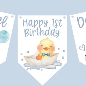 May include: A banner with three pennants featuring a watercolor illustration. The pennants display a yellow duck with a blue bow tie and a balloon. The text "Daniel" and "Happy 1st Birthday" are also present.