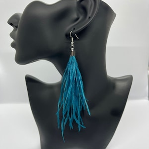 May include: A pair of long, teal feather earrings. The earrings are displayed on a black mannequin head. The feathers are a vibrant teal colour and hang down from silver-coloured hooks.