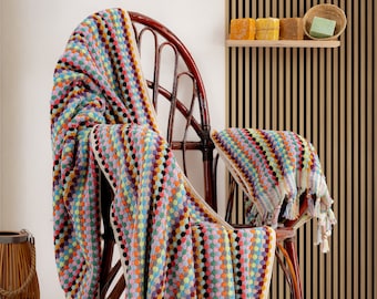 May include: A colorful striped towel with a waffle texture is draped over a wicker chair. The towel has a fringe border and is made of cotton.