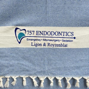 Op de afbeelding: Een wit-blauwe gestreepte handdoek met een franje rand. De handdoek heeft een logo met de tekst "757 Endodontics" met daaronder de tekst "Emergency. Microsurgery. Sedation" en "Ligon & Royzenblat".