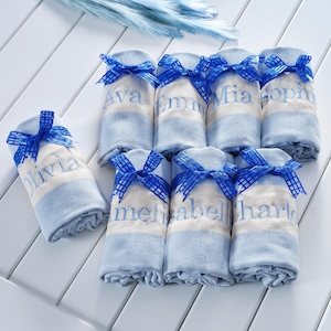 May include: A collection of seven rolled towels featuring light blue and cream geometric patterns. Each towel is tied with a blue checkered ribbon and embroidered with a unique name including Ava, Emma, Mia, Sophia, Olivia, Amelia, Isabella, and Charlotte.