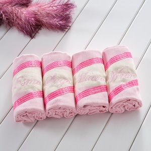 May include: Four rolled towels featuring a light pink and cream diamond-patterned weave. Each towel is accented with two horizontal bright pink textured stripes and embroidered cursive text in pink thread, displaying the names Esme, Sage, Wave, and Wren.
