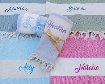 Op de afbeelding: Vijf gepersonaliseerde Turkse handdoeken met franjes. De handdoeken zijn gevouwen en in een rij gerangschikt. De handdoeken zijn in de kleuren blauw, groen, paars, lichtblauw en roze. De namen Maddox, Brianna, Susy, Heather, Ally en Natalie zijn geborduurd op de handdoeken.