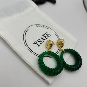 Peut inclure: Une paire de boucles d'oreilles créoles en jade vert avec des accents dorés. Les boucles d'oreilles sont suspendues à un sac en tissu blanc avec le texte "SCAUX YSAEE" imprimé dessus.