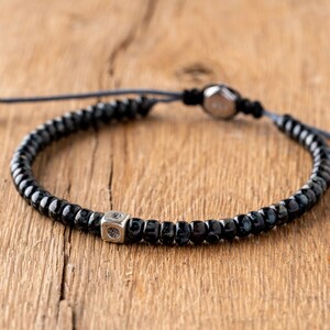 Op de afbeelding: Zwart kralenarmband met een zilveren dobbelsteenbedel. De armband heeft kleine, donkere kralen geregen op een donker koord. De dobbelsteenbedel is vierkant met geëtste details. Op een houten ondergrond.