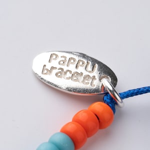 Op de afbeelding: Een close-up van een blauw en oranje kralenarmband met een zilveren bedel waarop "Pappu bracelet" staat.