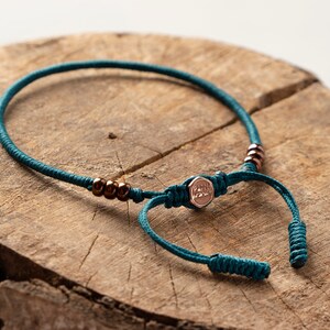 Op de afbeelding: Tealblauwe gevlochten koord armband met een roségouden bedel en kleine koperen kralen. De bedel heeft het woord "Faith" gegraveerd.