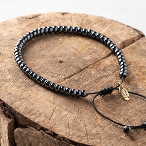 Op de afbeelding: Een zwarte kralenarmband met een schuifknoopsluiting. De armband is gemaakt van kleine, ronde, donkergrijze kralen geregen op een zwart koord. Een klein goudkleurig label met tekst hangt aan het koord. Op een houten oppervlak.