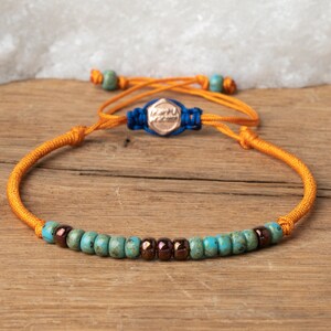 Op de afbeelding: Een gele koordarmband met blauwe en turquoise kralen en een roségouden bedel met het woord "Happy" erop.
