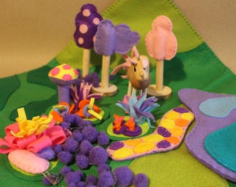 Puede incluir: Un conjunto de juego de fieltro con setas moradas, rosas y amarillas, una mariposa morada, una flor rosa y un camino morado y azul. El conjunto está sobre un fondo de fieltro verde.