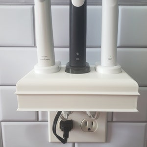 May include: White charging station with three toothbrush holders. The station has a white cord plugged into a standard electrical outlet. The toothbrushes are white, black, and gray.