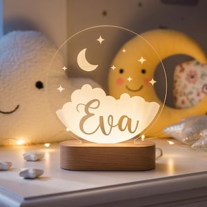 May include: A personalized night light with the name "Eva" etched on a cloud-shaped acrylic panel. The light sits on a wooden base and features a crescent moon and stars design. The image also includes a cloud-shaped pillow and a smiling moon.
