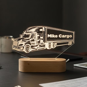 May include: A lighted truck-shaped desk lamp with the text "Mike Cargo" on the trailer. The lamp is made of clear acrylic and sits on a wooden base. The lamp is dimmable and has a cool-warm white light setting. Made in the USA.