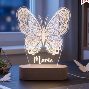 May include: A decorative butterfly-shaped LED lamp with the name "Marie" etched on it. The lamp sits on a wooden base and is surrounded by other butterfly decorations. The lamp is dimmable and offers cool-warm white light.