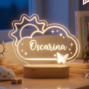 May include: A personalized night light with the name "Oscariana" in a cloud shape, featuring a sun, stars, and a butterfly. The lamp sits on a wooden base and is dimmable. The light offers cool-warm white options.