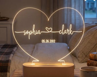 Puede incluir: Luz de noche LED en forma de corazón con los nombres "Sophia" y "Clark" y la fecha "05.26.2012" iluminados en blanco cálido. La luz se apoya en una base de madera y es regulable. Fabricado en EE. UU.