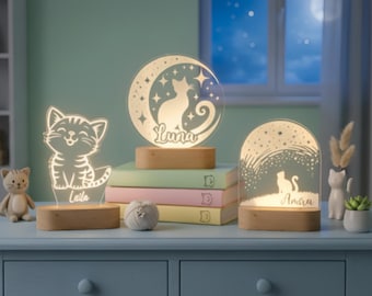 Könnte beinhalten: Drei beleuchtete Nachtlichter mit Katzenmotiven auf einem blauen Tisch. Eines zeigt eine Katzensilhouette, ein anderes eine Katze auf einem Mond und das dritte eine Katze unter einem Sternenhimmel. Jedes Licht steht auf einem Holzsockel. Die Lichter sind dimmbar und bieten kalt-warmweißes Licht.