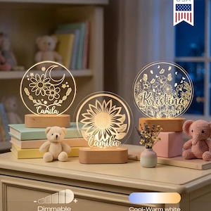 May include: Three illuminated personalized night lights with floral designs and names, set on wooden bases. One features a sunflower, another a moon and flowers, and the third a floral pattern. A small teddy bear and a pink elephant are also on the table.